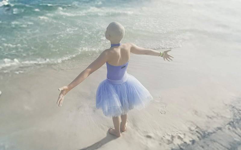 a child in a ballet outfit with no hair. arms extended and facing the ocean