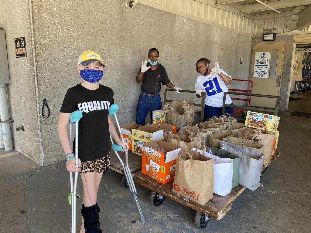 A child with crutches in a black t-shirt that says equality standing in front of a palette full of bags of food